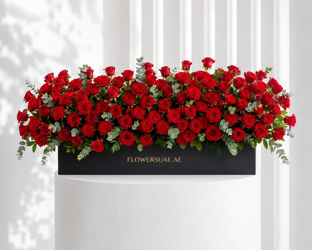 Arrangement of red roses with greenery on a black box labeled 'FLOWERSUAE.AE' against a white curtain background.