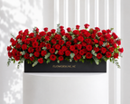 Arrangement of red roses with greenery on a black box labeled 'FLOWERSUAE.AE' against a white curtain background.