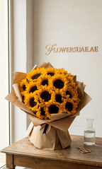 Large bouquet of fresh sunflowers wrapped in paper on a wooden table beside a glass vase and scissors